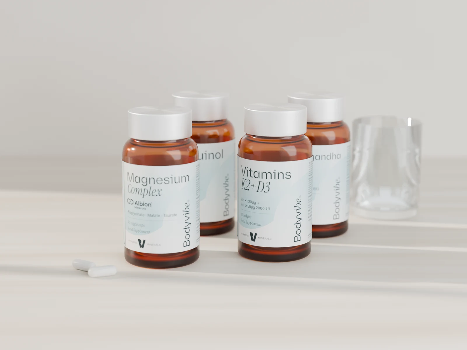 Magnesium Complex, Vitamins K2+D3, Ubiquinol CoQ10 y Ashwagandha Complex en sus botes de vidrio ámbar sobre piedra, con luz de ventana y un vaso de agua